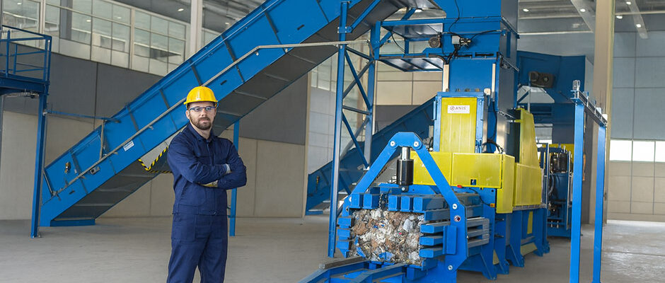 Vzdrževanje balirke v industrijskem obratu z delavcem v zaščitni opremi poleg avtomatizirane stiskalnice za odpadke.